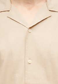 Κοντινό πλάνο μπεζ πουκάμισου με γιακά με χαραγή, με δύο κουμπιά κλεισμένα στο μπροστινό μέρος.