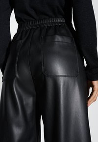 Schwarze Lederhosen mit glatter Textur, ausgestattet mit einem elastischen Taillenbund und zwei Gesäßtaschen. Das Design ist weit geschnitten und bietet einen lockeren Sitz.