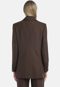 Persona con cabello largo y rubio vista desde atrás, vistiendo un blazer oscuro ajustado con puños abotonados y pantalones a juego.