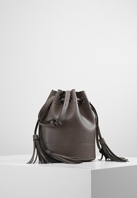 Harmaa nahkainen säkylaukku, jossa on nyörikiristys, tupsuaksentteja ja sileä pinta, lepää vaalean värisellä pinnalla.