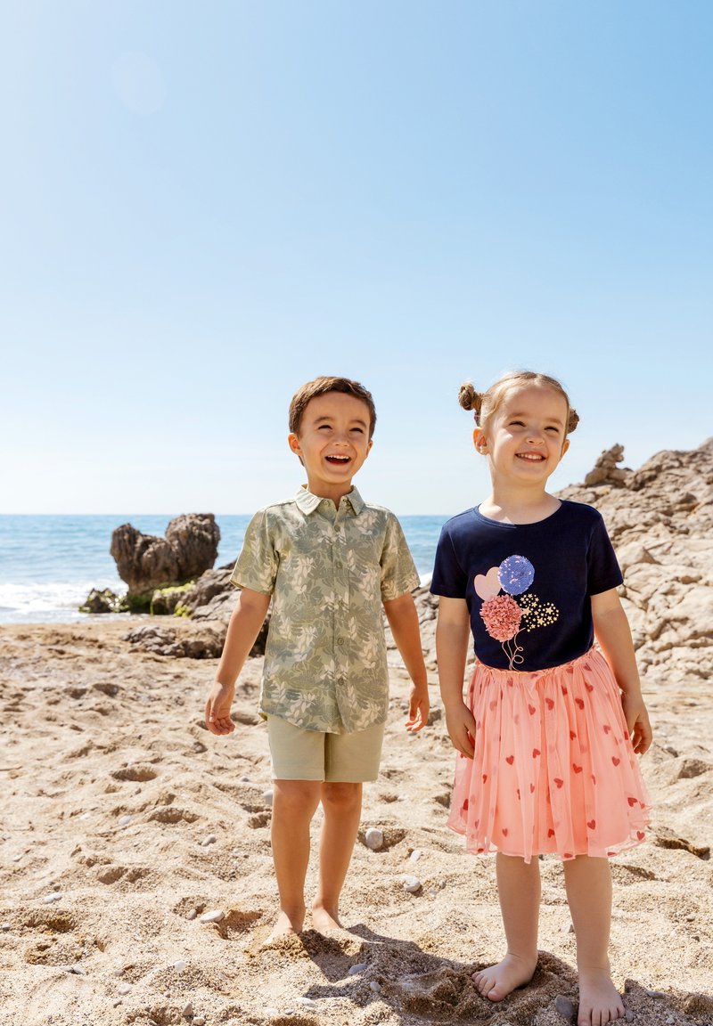 To børn står på en sandet strand. Drengen har en lysegrøn skjorte med blomsterprint og beige shorts på. Pigen har en marineblå skjorte med ballonmotiver og et lyserødt skørt med hjerter.