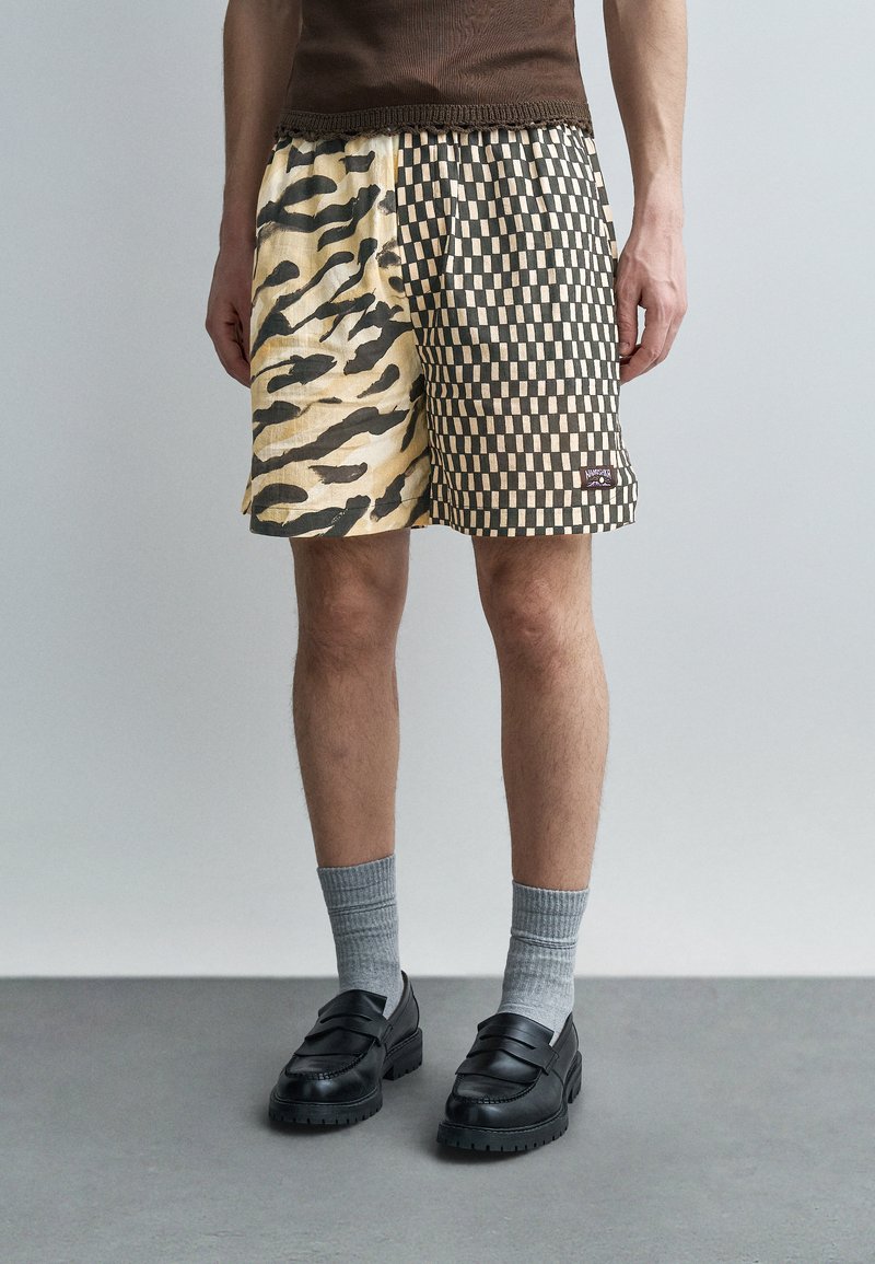 Homme portant un short à design fendu : côté gauche beige avec imprimé animalier noir, côté droit noir avec motif damier beige, chaussures foncées et chaussettes grises.