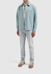 Lichtblauw, knoopshirt met twee voorzakken, gedragen over een crèmekleurig shirt, gecombineerd met lichte spijkerbroek en bruine sneakers met veters.