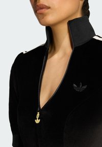 Crna baršunasta jakna na zip s visokim ovratnikom, koja sadrži zlatni zip i izvezeni logo na prsima, ističući glatku teksturu.