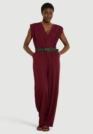 Donna che indossa una tuta bordeaux senza maniche con scollo a V e cintura nera, in piedi con le mani nelle tasche e calzando sandali.