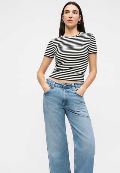 Γυναίκα με μακριά σκούρα μαλλιά που φοράει ασημένια σκουλαρίκια σε σχήμα κρίκου, μαύρο-άσπρο ριγέ μπλουζάκι με κοντά μανίκια και ανοιχτό μπλε τζιν ψηλόμεσο.