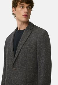 Grau gewebter Blazer mit strukturierter Oberfläche, mit Reverskragen, zwei vorderen Taschen und einem Knopfverschluss, getragen über einem schwarzen Oberteil.