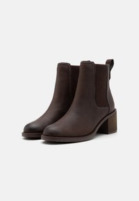 Botas de tobillo de cuero marrón con paneles laterales elásticos, punteras redondeadas y un tacón de madera apilada. Una tela texturizada acentúa el diseño.