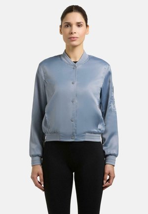 Kobieta ubrana w błękitną satynową kurtkę typu bomber z zatrzaskami i czarne spodnie, stojąca na jednolitym białym tle.