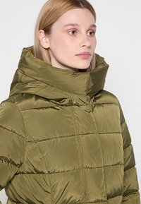 Chaqueta acolchada de color verde oliva con un alto cuello, diseño acolchado y textura brillante. Presenta un cierre frontal y un detalle de costura minimalista.