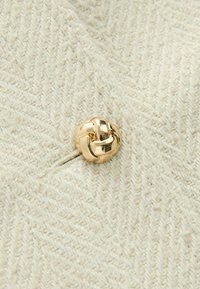 Crèmekleurige visgraatstof met een glanzende gouden knoop in de vorm van een knoop erop genaaid.