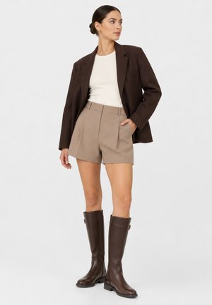 Kvinde iført en mørkebrun blazer, hvid top, beige shorts og mørkebrune læderstøvler op til knæet står foran en ensfarvet baggrund.