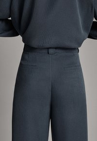 Personne portant un pantalon taille haute texturé foncé avec des poches passepoilées à l’arrière et un haut ample assorti, montré de la taille jusqu’à mi-cuisse.