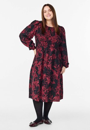 Glunderende vrouw met steil bruin haar, gekleed in een zwart en rood bloemen midi-jurk, zwarte panty’s en bordeauxrode ballerina's met gespen.