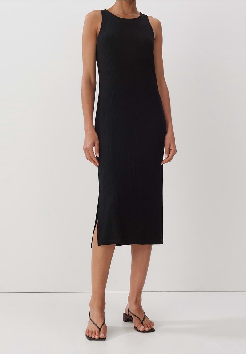 Kvinna i en ärmlös svart midiklänning med sidoslits och svarta sandaler med låg klack och remmar, stående mot en neutral bakgrund.