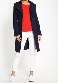 Kvinna som har på sig en marinblå trenchcoat över en röd topp, vita uppkavlade byxor och vita slip-on skor, stående mot en enkel vit bakgrund.