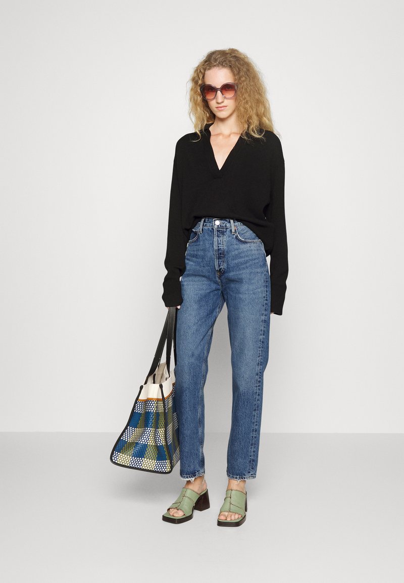 Un top noir à manches longues en v, un jean en denim bleu taille haute, des sandales à plateformes vertes et un sac fourre-tout tissé multicolore avec des poignées noires.