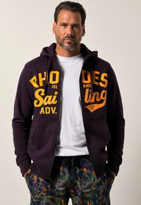 Mørkelilla zip-up højhalstrøje med store gule bogstaver "RHODES SAILING ADV." på forsiden, parret med mønstrede blomsterbukser.