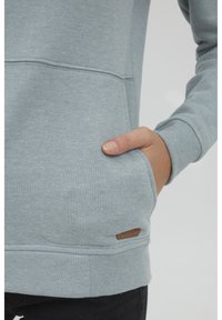 Felpa grigio-blu in morbido tessuto, con tasca a marsupio e polsini a costine. Accento del logo marrone vicino all'orlo.