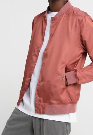 Persona che indossa una giacca bomber in satin color ruggine con bottoni a scatto, una maglietta bianca e pantaloni grigi, con una mano nella tasca della giacca.