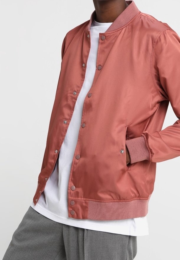 Person wearing a rust-colored satin bomber jacket with snap buttons, a white t-shirt, and gray trousers, hand in jacket pocket.