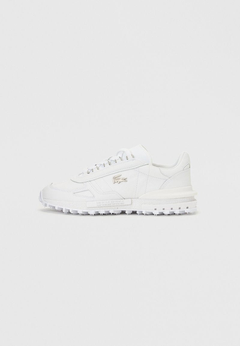 Baskets basses blanches avec semelle texturée, fermeture à lacets à l'avant et logo crocodile argenté sur le côté, sur fond gris uni.
