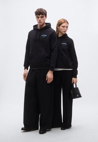 Μαύρα φούτερ με κουκούλα, εσωτερικές τσέπες και κορδόνια, με πολύχρωμα κείμενα. Τα μοντέλα φορούν φαρδιά μαύρα παντελόνια και κρατούν μικρή μαύρη τσάντα.