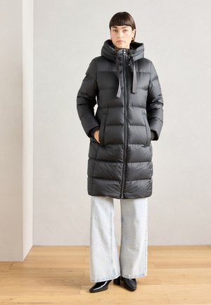 Manteau mi-long noir rembourré avec col montant, fermeture éclair et cordons de serrage, associé à un jean en denim bleu clair à jambes larges et des bottes noires.