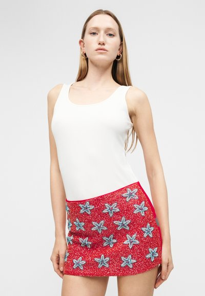 Γυναίκα που φορά λευκό αμάνικο τοπ και κοντή κόκκινη φούστα με μπλε και λευκά σχέδια αστερίσκων, στέκεται μπροστά σε απλό φόντο.