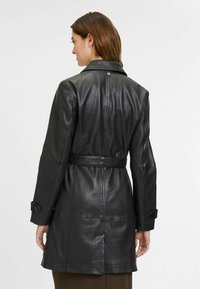 Cappotto di pelle nera con vita a scorrimento, maniche lunghe e dettagli a bottoni sui polsini. Texture liscia con una vestibilità sartoriale.