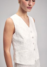 Wit gebloemd vest met V-hals, vijf knopen en een zijzak. Het materiaal heeft een gestructureerd patroon.