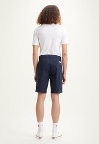 Shorts bleu marine avec une texture lisse, deux poches arrière et un patch de marque. Portés avec une chemise blanche, des chaussettes blanches et des chaussures blanches.