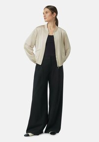 Hellbeige Bomberjacke mit Rundhalsausschnitt, elastischen Bündchen und zwei Seitentaschen, kombiniert mit schwarzen, weiter geschnittenen Hosen und spitzen Schuhen.