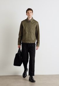 Olivengrønn zip-up jakke med krage, kombinert med svarte bukser og svarte sko. Modell som bærer en svart veske, står mot en hvit bakgrunn.