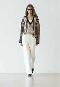 Maglione a righe lavorato a maglia in beige e blu navy, design a scollo a V, abbinato a pantaloni bianchi a gamba larga e scarpe nere. La modella indossa anche occhiali da sole.