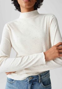 Cremefarbener, gerippter, langärmeliger Rollkragenpullover mit schwarzen Sprenkeln und Rüschensäumen, getragen mit blauen Jeans.