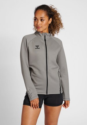 Kobieta o kręconych włosach, ubrana w szarą sportową kurtkę na zamek i czarne szorty, stojąca na jednolitym białym tle.