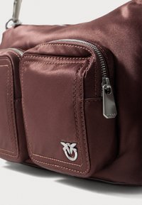Burgundowa satynowa torba z dwiema kieszeniami na zamek, srebrnymi akcentami i logo. Charakteryzuje się gładką teksturą i odsłoniętym szwem.