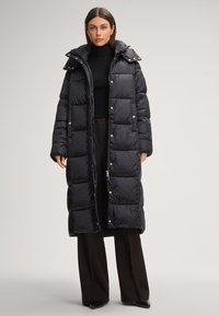 Lungo cappotto imbottito nero con motivo trapuntato, bottoni metallici e cappuccio staccabile. Indossato sopra un dolcevita nero e pantaloni scuri.