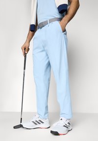 Jasnoniebieskie golfowe spodnie z płaskim przodem, z szarym paskiem, w połączeniu z białymi butami golfowymi i czarnym putterem.