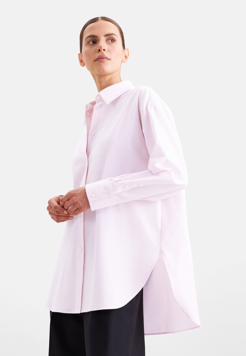 Femme aux cheveux tirés en arrière, portant une chemise rose clair oversize boutonnée et un pantalon noir sur un fond clair uni.
