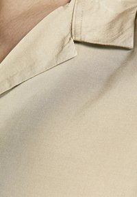 Tejido de color beige claro con una textura suave, que presenta un collar puntiagudo y un ligero brillo. Un acercamiento revela los finos detalles del tejido.