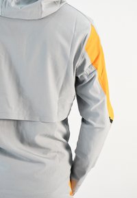 Chaqueta con capucha gris de diseño corto, con acentos naranjas en los hombros y un tejido suave y texturizado.