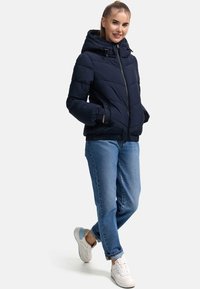 Veste matelassée bleu marine avec capuche, fermeture éclair et poignets élastiques, associée à un jean bleu clair et des baskets blanches.