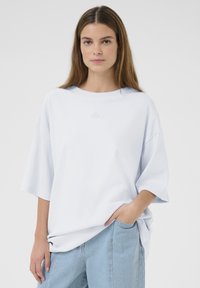 Oversized lichtblauwe katoenen t-shirt met verlaagde schouders, ronde halslijn en subtiel geborduurd logo op de voorkant. Gecombineerd met lichtblauwe jeans.