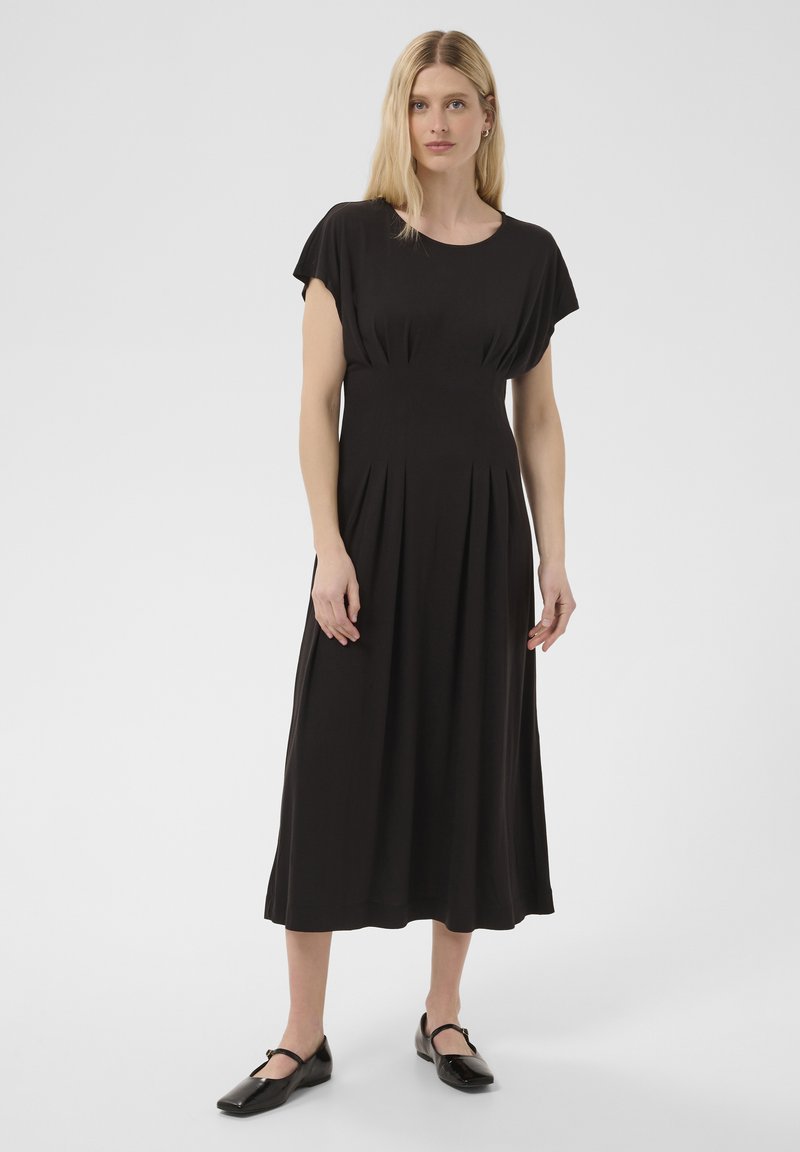 Femme blonde portant une robe midi noire à manches courtes et des ballerines Mary Jane noires, debout sur un fond blanc uni.