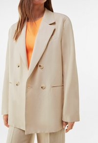 Veste beige croisée à gros boutons, col à revers cranté et poches latérales, conçue dans un tissu lisse. Portée sur un haut orange.
