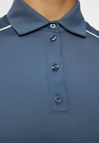 Lähikuva henkilöstä, jolla on päällään sininen polo-paita, jossa on neljä sinistä nappia ja valkoinen raitadetaili olkapäällä.