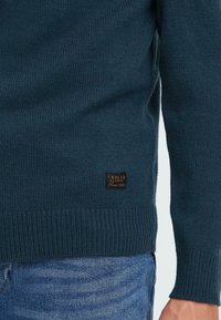 Mørk teal strikket genser med ribbede mansjetter og hem, med et brunt skinnmerke som har "S.OLID Denmark Siden 1999" på siden.