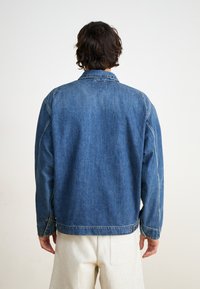 Denimbiks jacket vidējā zilā krāsā, ar klasisku apkakli, garām piedurknēm un redzamām šuvēm ar metāla pogu akcentiem.